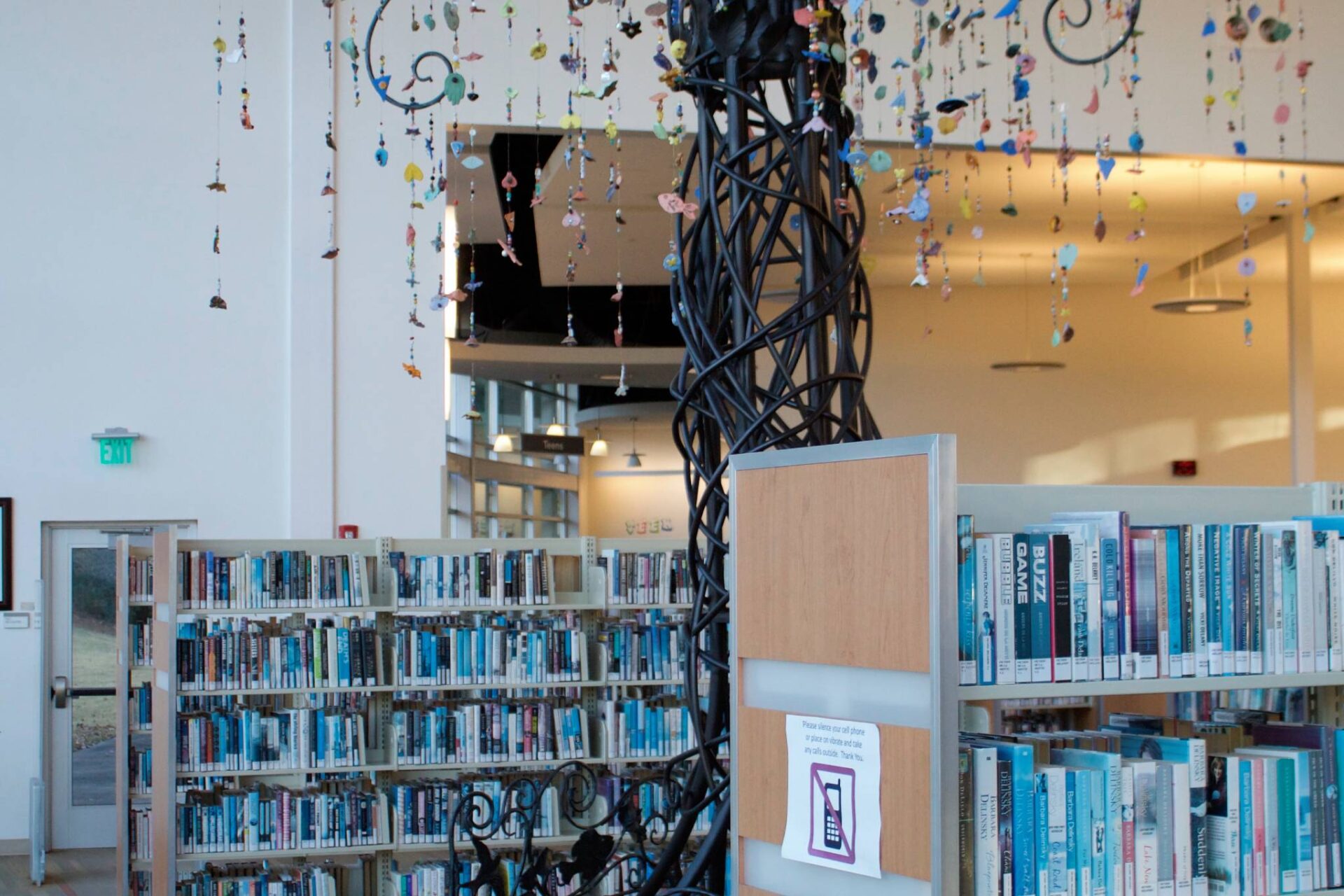 Artistic-metal-tree-sculpture-with-colorful-hanging-elements-in-a-library-surrounded-by-bookshelves-scaled