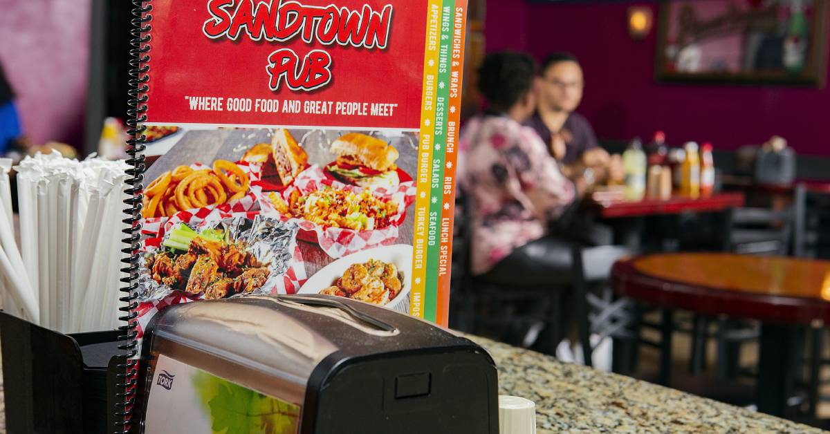 Menu of a pub featuring food images, with a napkin dispenser in front and tables in the background.