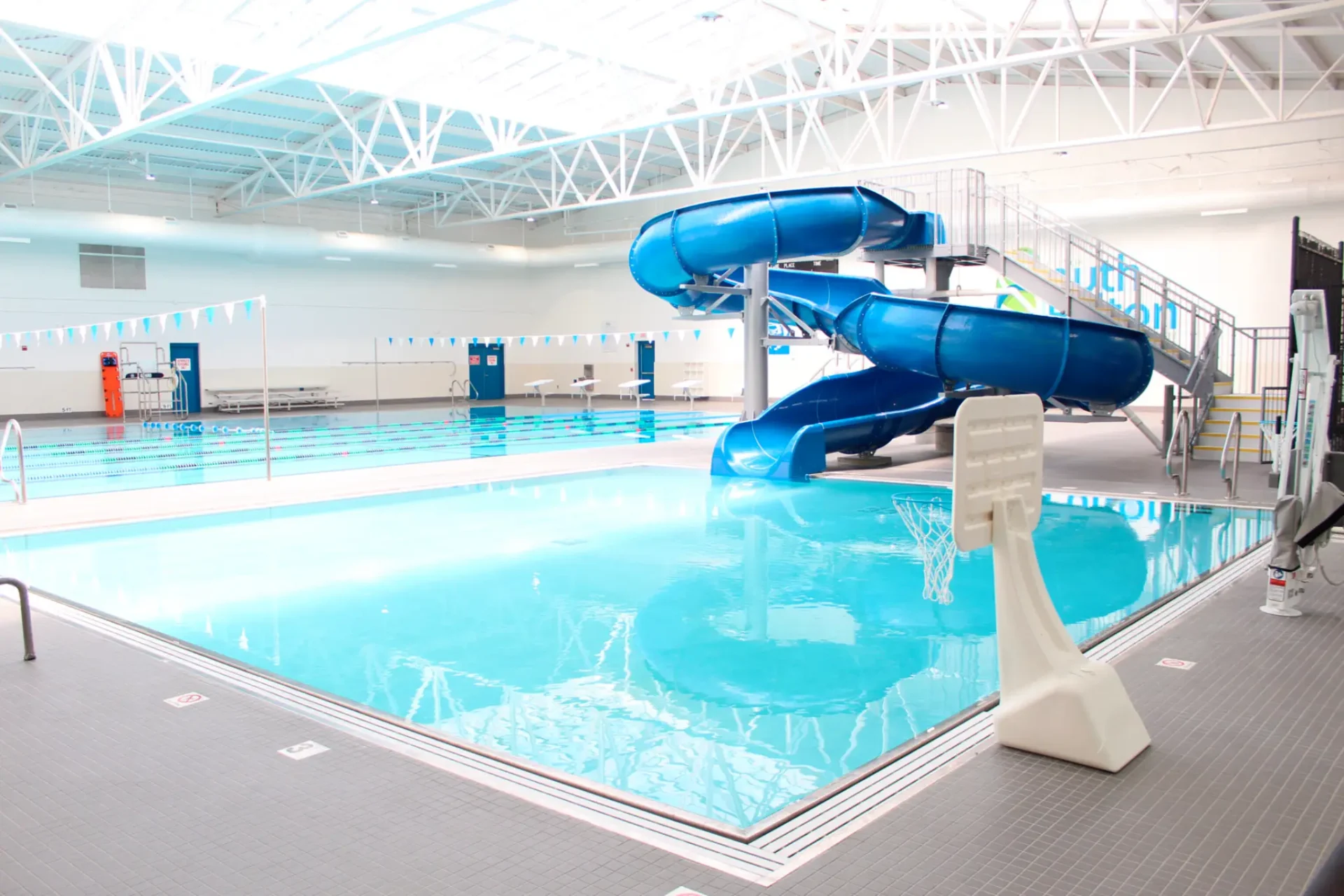 Indoor swimming pool featuring a blue waterslide, diving boards, lifeguard station, and basketball hoop at the edge of the water